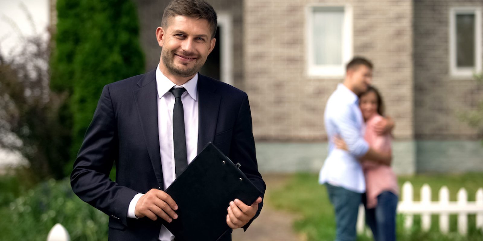 Smiling broker standing with documents, happy family hugging near their new home