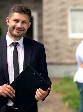 Smiling broker standing with documents, happy family hugging near their new home